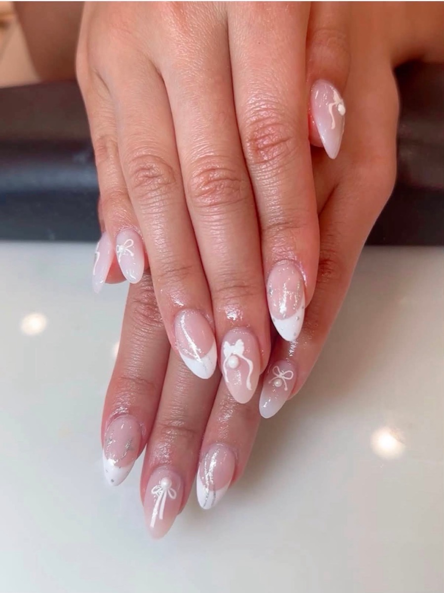 Nude almond nails finished with a hand-painted white bow and pearl accents at Lumi Studio